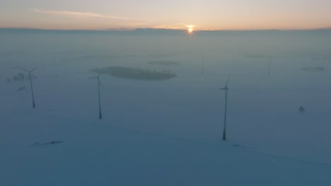 Aerial-view-of-wind-turbines-generating-renewable-energy-in-the-wind-farm,-snow-filled-countryside-landscape-with-fog,-sunny-winter-evening-with-golden-hour-light,-wide-drone-shot-moving-forward