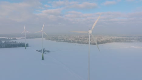 Aerial-view-of-wind-turbines-generating-renewable-energy-in-the-wind-farm,-snow-filled-countryside-landscape-with-fog,-sunny-winter-evening-with-golden-hour-light,-wide-drone-shot-moving-bacward