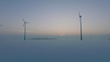 Aerial-view-of-wind-turbines-generating-renewable-energy-in-the-wind-farm,-snow-filled-countryside-landscape-with-fog,-sunny-winter-evening,-golden-hour-light,-establishing-drone-shot-moving-backward