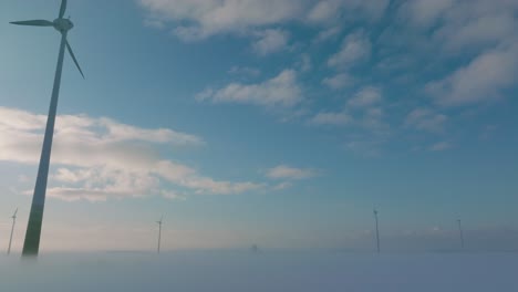 Aerial-establishing-view-of-wind-turbines-generating-renewable-energy-in-the-wind-farm,-snow-filled-countryside-landscape-with-fog,-sunny-winter-evening-golden-hour,-wide-drone-shot-moving-forward-low