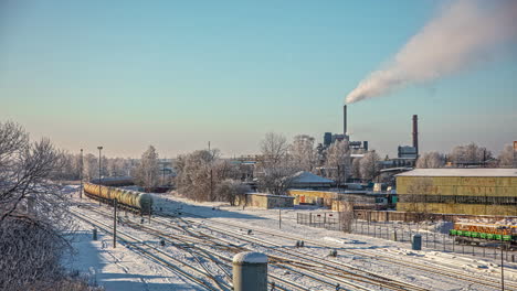 The-industrial-part-of-a-town-in-winter-with-a-tanker-car-train-running-by-as-steam-come-out-of-a-factory-smokestack---time-lapse