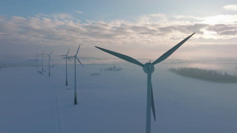Aerial-establishing-view-of-wind-turbines-generating-renewable-energy-in-the-wind-farm,-snow-filled-countryside-landscape-with-fog,-sunny-winter-day,-wide-orbiting-drone-shot