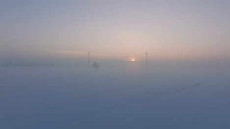 Aerial-view-of-wind-turbines-generating-renewable-energy-in-the-wind-farm,-snow-filled-countryside-landscape-with-fog,-sunny-winter-evening-with-golden-hour-light,-wide-drone-shot-moving-forward-low