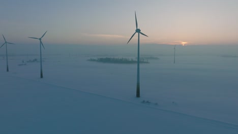 Aerial-view-of-wind-turbines-generating-renewable-energy-in-the-wind-farm,-snow-filled-countryside-landscape-with-fog,-sunny-winter-evening-with-golden-hour-light,-ascending-drone-shot-moving-forward