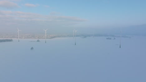 Establishing-aerial-view-of-wind-turbines-generating-renewable-energy-in-the-wind-farm,-snow-filled-countryside-landscape-with-fog,-sunny-winter-day,-wide-drone-shot-moving-forward