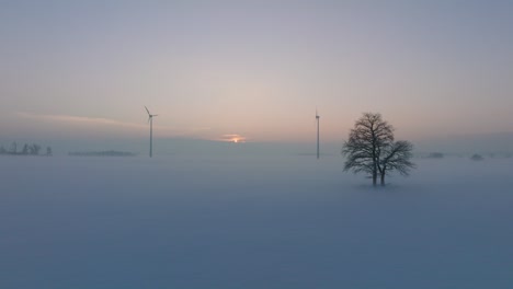 Aerial-view-of-wind-turbines-generating-renewable-energy-in-the-wind-farm,-snow-filled-countryside-landscape-with-fog,-sunny-winter-evening-with-golden-hour-light,-wide-drone-shot-moving-forward-low