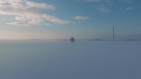 Aerial-establishing-view-of-wind-turbines-generating-renewable-energy-in-the-wind-farm,-snow-filled-countryside-landscape-with-fog,-sunny-winter-evening,-wide-drone-shot-moving-forward-low