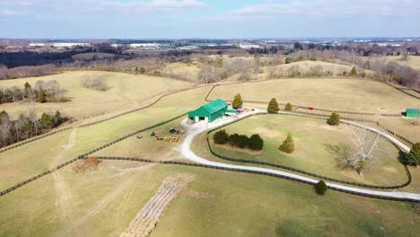 Aerial-reveal-of-a-large-green-horse-barn-in-the-middle-of-the-bluegrass-state-Frankfort-Kentucky-with-grand-blue-sky-vistas-and-gorgeous-evergreen-trees-and-dynamic-shadows