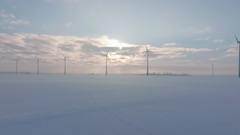 Establishing-aerial-view-of-wind-turbines-generating-renewable-energy-in-the-wind-farm,-snow-filled-countryside-landscape-with-fog,-sunny-winter-day,-wide-drone-shot-moving-forward-low