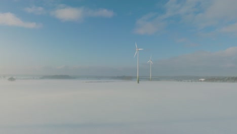 Aerial-establishing-view-of-wind-turbines-generating-renewable-energy-in-the-wind-farm,-snow-filled-countryside-landscape-with-fog,-sunny-winter-day,-wide-ascending-drone-shot-moving-forward