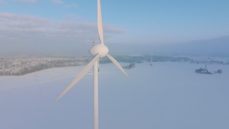 Aerial-view-of-wind-turbine-generating-renewable-energy-in-the-wind-farm,-snow-filled-countryside-landscape-with-fog,-sunny-winter-evening-with-golden-hour-light,-wide-drone-shot-moving-backward