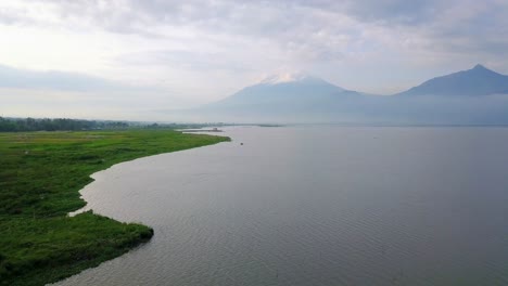 Aerial-view-of-huge-lake-and-green-meadow-on-the-lakeside-with-mountain-on-the-background---Rawa-Pening-Lake,-Indonesia