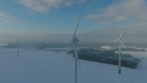 Aerial-view-of-wind-turbines-generating-renewable-energy-in-the-wind-farm,-snow-filled-countryside-landscape-with-fog,-sunny-winter-day-with-some-clouds,-wide-orbiting-drone-shot