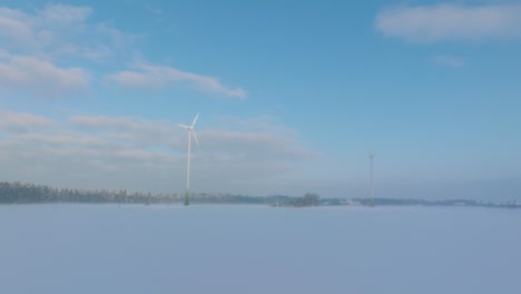 Aerial-view-of-wind-turbines-generating-renewable-energy-in-the-wind-farm,-snow-filled-countryside-landscape-with-fog,-sunny-winter-day,-wide-drone-shot-moving-forward