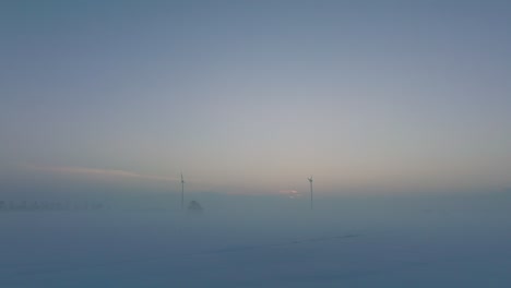 Aerial-view-of-wind-turbines-generating-renewable-energy-in-the-wind-farm,-snow-filled-countryside-landscape-with-fog,-sunny-winter-evening-with-golden-hour-light,-low-drone-shot-moving-backward