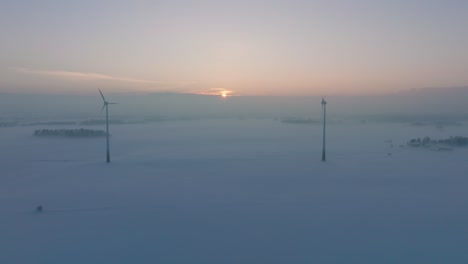 Aerial-view-of-wind-turbines-generating-renewable-energy-in-the-wind-farm,-snow-filled-countryside-landscape-with-fog,-sunny-winter-evening-with-golden-hour-light,-wide-drone-shot-moving-backward