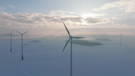 Aerial-establishing-view-of-wind-turbines-generating-renewable-energy-in-the-wind-farm,-snow-filled-countryside-landscape-with-fog,-sunny-winter-evening-golden-hour,-wide-drone-shot-moving-forward