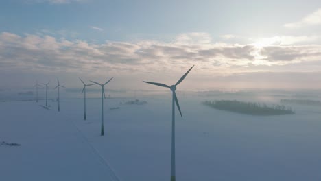 Aerial-establishing-view-of-wind-turbines-generating-renewable-energy-in-the-wind-farm,-snow-filled-countryside-landscape-with-fog,-sunny-winter-evening,-golden-hour-light,-wide-drone-orbiting-shot