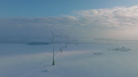 Aerial-view-of-wind-turbines-generating-renewable-energy-in-the-wind-farm,-snow-filled-countryside-landscape-with-fog,-sunny-winter-evening-with-golden-hour-light,-wide-drone-shot