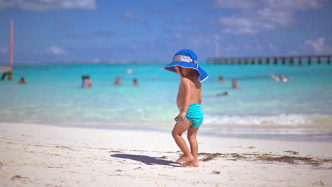 Little-baby-boy-with-turquoise-diapers-and-a-blue-hat-standing-on-the-beach-watching-the-sea-holding-a-blue-car-in-Cancun-Mexico
