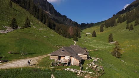 Aerial,-male-backpacker-walking-towards-rural-cabin-by-the-mountains-during-summer