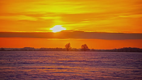 Bright-yellow-to-orange-sunset-as-the-sun-descends-over-the-winter,-snowy-horizon---time-lapse