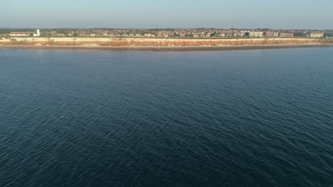 High-Aerial-Drone-Shot-Flying-Away-From-Old-Hunstanton-White-and-Orange-Cliffs-with-Lighthouse-on-Top-Over-Calm-Water-with-Small-Waves-in-North-Norfolk-UK