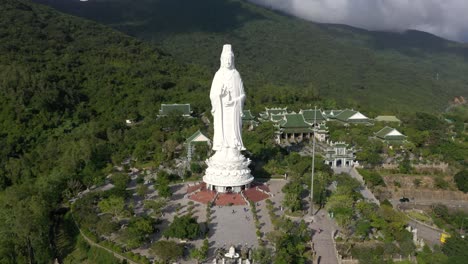 Close-aerial-circling-around-tall-Lady-Buddha-statue-and-temples-with-huge-mountains-and-ocean-in-Da-Nang,-Vietnam