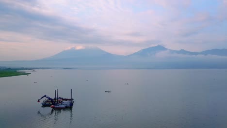 Flyover-view-of-Indonesia-lake-with-digger-boat-and-mountains-in-background