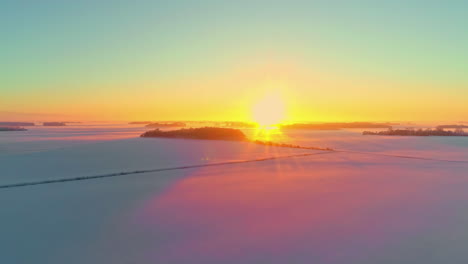 Aerial-drone-flying-over-snow-covered-landscape-with-snow-covered-farmland-at-sunrise-along-bright-yellow-sky