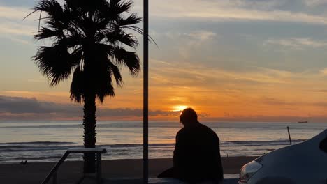 Silhouette-of-young-adult-man-smoking-a-cigarette-in-sunset-and-with-some-surfers-enjoying-the-sea