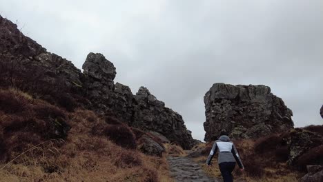 Mujer-Caminando-Por-Un-Camino-De-Piedra-Que-Sube-Las-Colinas-Con-Hierba-Y-Un-Entorno-Rocoso-En-Islandia