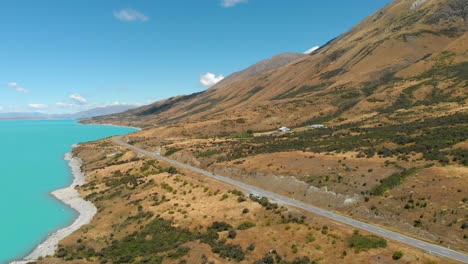Aerial-view-of-a-road-lakeside-to-Lake-Pukaki,-New-Zealand,-while-a-camper-is-driving-on
