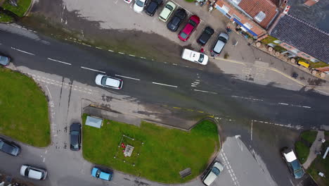 Topdown-Aerial-Drone-Shot-of-Silver-and-Red-Cars-and-White-Van-Driving-Through-Water-on-Flooded-Road-in-Small-UK-Village