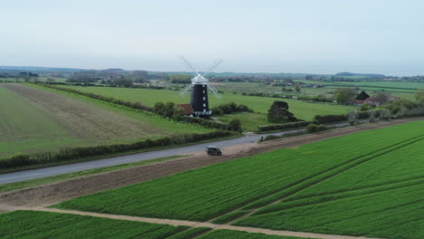 Rising-Aerial-Drone-Shot-Of-Rural-Old-19th-Century-Grade-II-Listed-Windmill-and-Green-Fields-with-Old-Landrover-Defender-Parked-on-Side-of-Road-Burnham-Overy-Staithe-North-Norfolk-UK