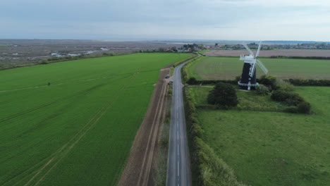 High-Aerial-Drone-Shot-Flying-Down-Road-with-Old-Windmill-with-Fields-and-Marshes-in-Shot-in-Burnham-Overy-Staithe-North-Norfolk-UK