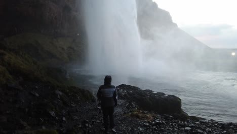 Mädchen,-Das-Unter-Wasserfall-Slowmotion-Aufnahme-Von-Unten-Nach-Oben-In-Der-Dunklen-Höhle-Islands-Steht