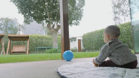 Niño-Jugando-Con-Una-Bola-Azul-Sentado-En-El-Patio-Trasero-De-Casa-Con-Vistas-Al-Jardín-A-La-Luz-Del-Día