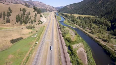 Arial-shot-of-train-driving-alongside-a-river-and-highwayon-USA's-westcoast-in-mountain-terrain