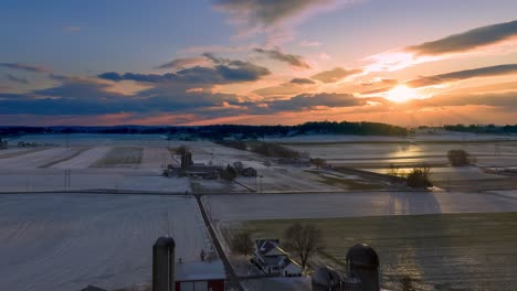 Aerial-hyperlapse-of-rolling-clouds-in-dramatic-sunset-above-rural-landscape,-sun-beams-reflect-in-frozen-fields,-traditional-farm-in-America,-winter-nature-time-lapse