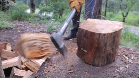 Winter-fuel---close-up-of-axe,-log,-man-at-work,-legs,-work-boots-and-work-gloves---rural-nature-bushland-behind-as-man-splits-log-with-axe---4K