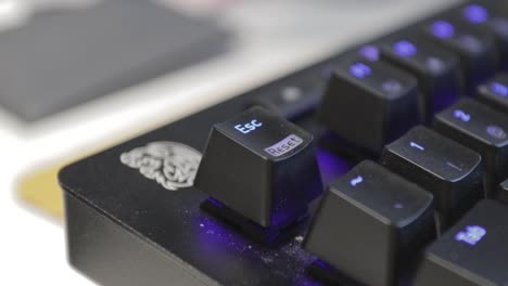 Drücken-Sie-Mit-Der-Hand-Eine-Einzelne-Taste-Auf-Einer-Leicht-Verstaubten,-Schwarzen-Tastatur-Mit-Blau-Beleuchteten-Tasten