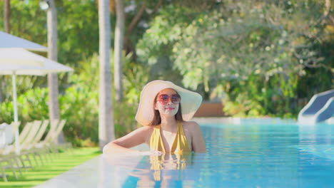 Smiling-tourist-standing-at-the-edge-of-a-pool,-wearing-big-sunglasses-and-a-floppy-sun-hat,-looking-at-the-camera-and-relaxing