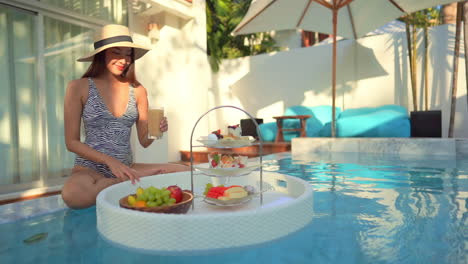 Young-woman-in-swimming-pool-eating-breakfast-from-floating-basket