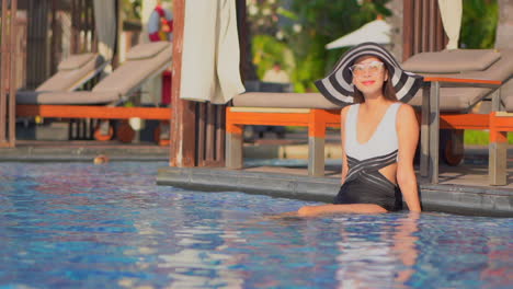 An-Asian-woman,-in-a-bathing-suit-and-huge-floppy-sun-hat,-sits-on-the-edge-of-a-resort-pool-with-feet-in-the-water-together