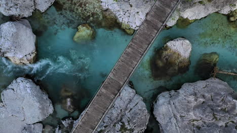 Top-down-view-of-a-wooden-footbridge-spanning-the-Soca-river-in-Slovenia-as-drone-rises-to-reveal-landscape