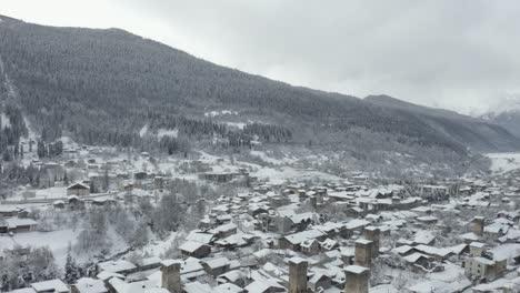 Weißer-Schnee-Auf-Den-Hausdächern-In-Mestia,-Svaneti,-Georgia-Im-Winter-Mit-Malerischem-Berg-Voller-Bäume-Im-Hintergrund---Luftaufnahme