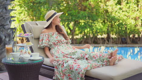 A-young-Asian-woman-in-a-long-loose-dress-and-straw-sun-hat-reclines-poolside-for-tea-time