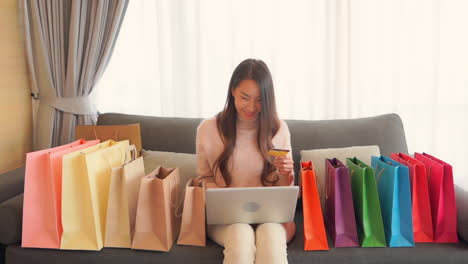 Attraktives-Asiatisches-Mädchen,-Umgeben-Von-Einkaufstüten,-Die-Ihre-Kreditkarte-Zum-Online-Shopping-Verwenden,-Geben-Sie-Karteninformationen-Auf-Dem-Laptop-Ein