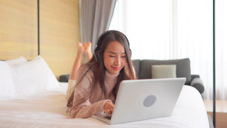 Slow-motion-of-relaxed-Asian-woman-using-her-laptop-computer-and-listening-to-the-music-lying-on-the-bed-with-her-legs-raised-at-home-wearing-casual-clothes